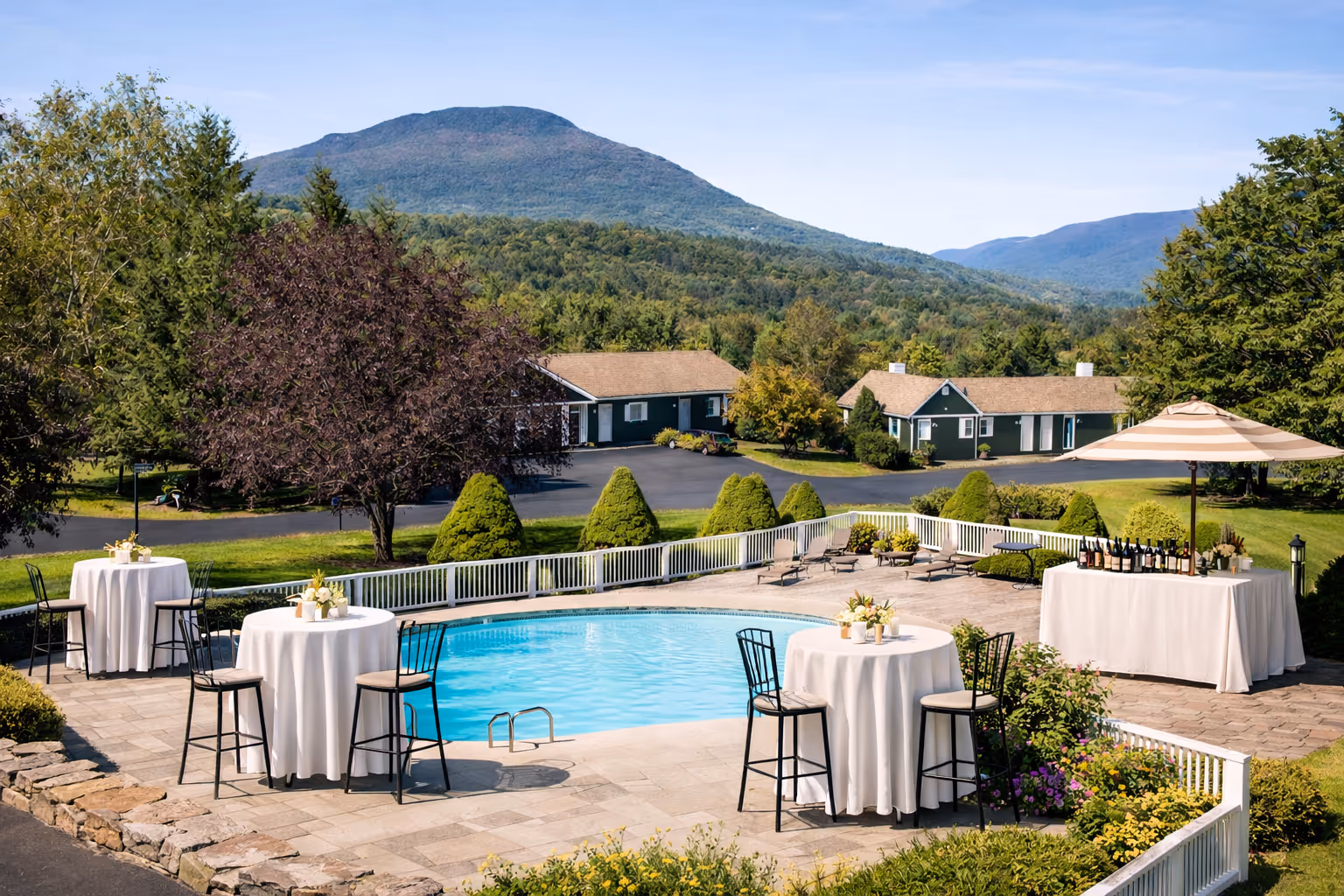 Outdoor pool area set for event with round tables, chairs, and a bar table under an umbrella, with green hills and houses in the background.