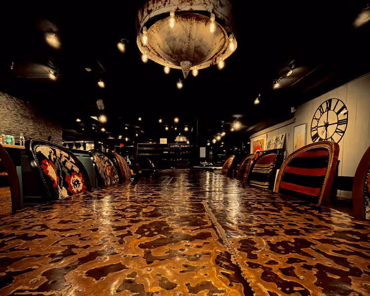 Long rustic wooden table surrounded by upholstered chairs with patterned fabrics under warm hanging lights in a dimly lit room with a large wall clock.