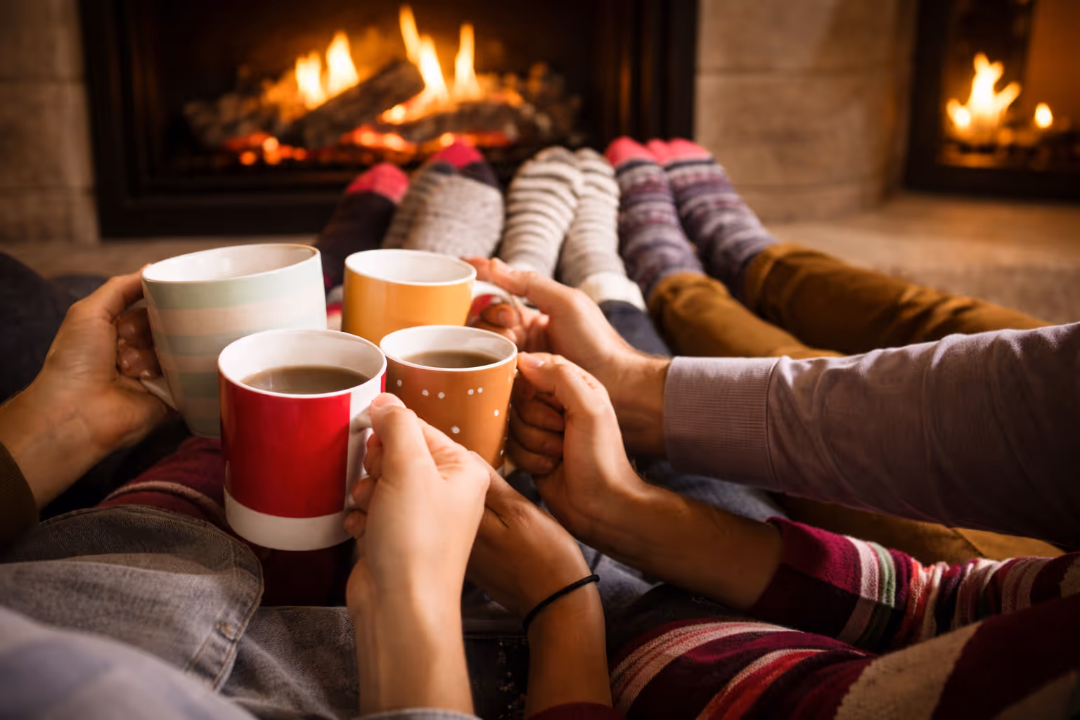 Group of people with colorful socks holding mugs of hot drinks near a cozy fireplace.