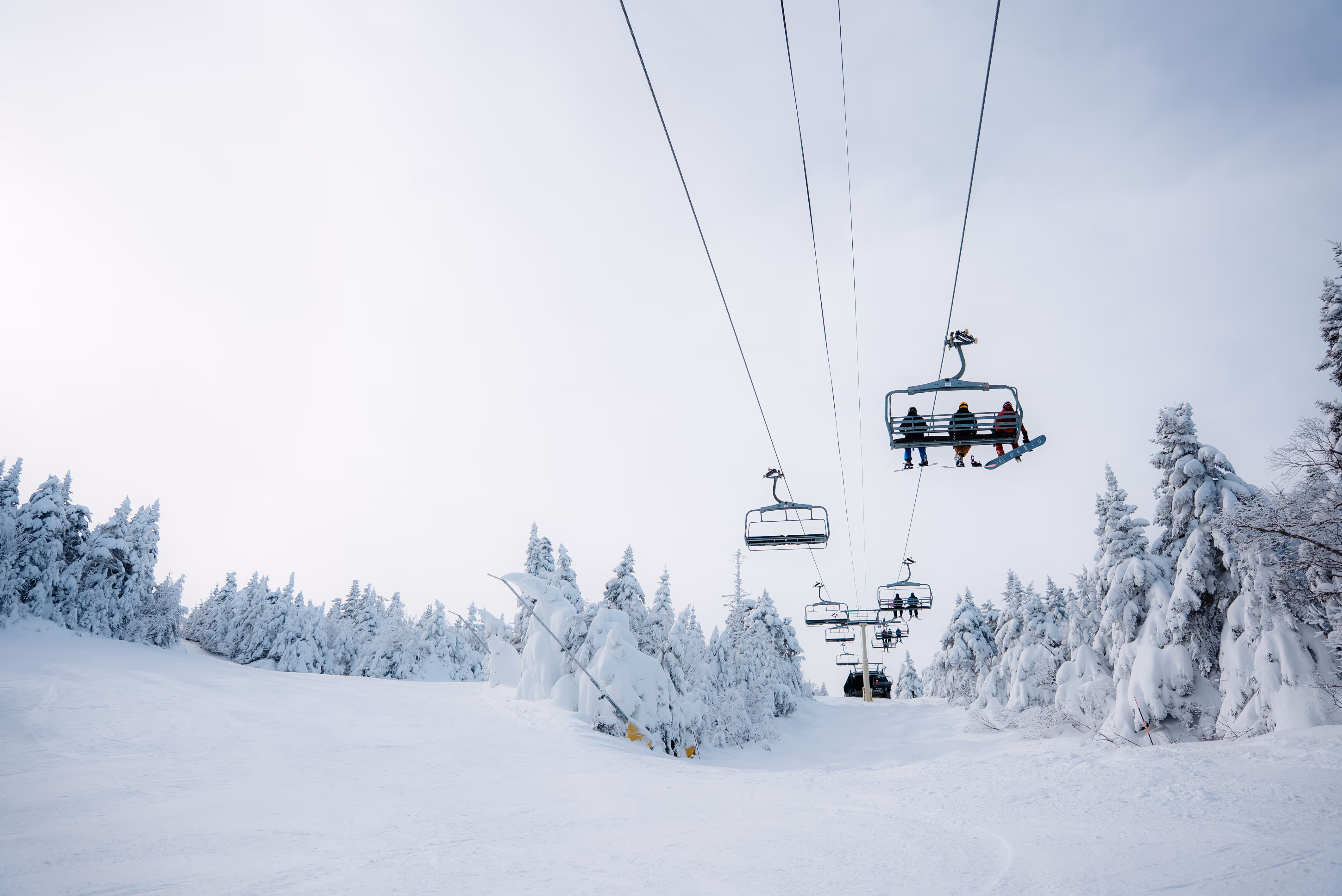 Ski lift carrying people over a snowy ski slope surrounded by snow-covered trees under a cloudy sky.