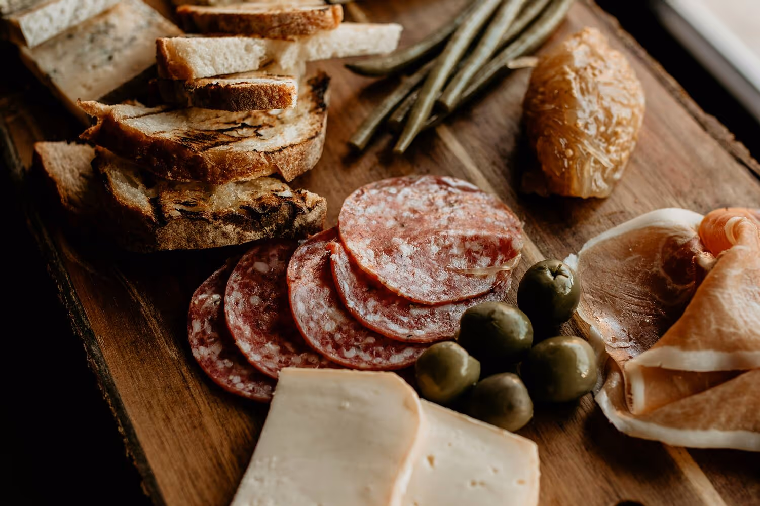 Wooden board with sliced bread, salami, green olives, cheese, ham, and pickled vegetables.
