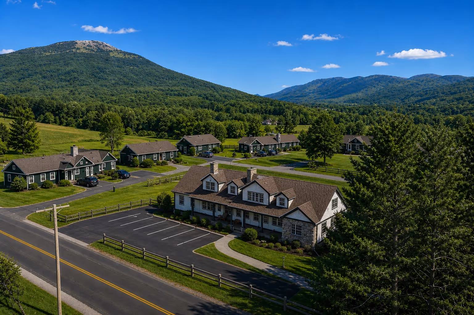 Cluster of green houses and a larger white building with parking, set against forested mountains under blue sky.