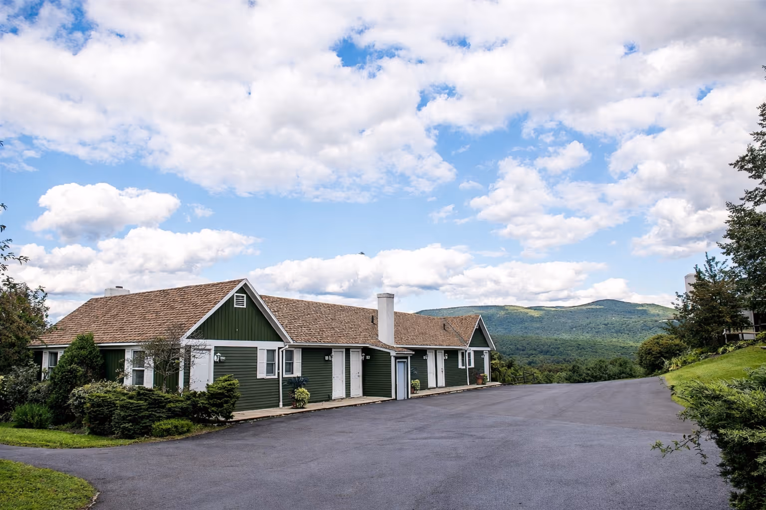 Single-story green building with white doors and shutters against a backdrop of trees and mountains.