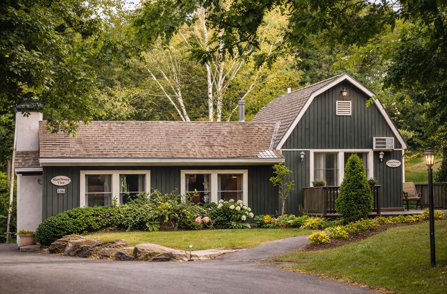 Green house with brown roof and landscaped garden with trees and flowering bushes in front.