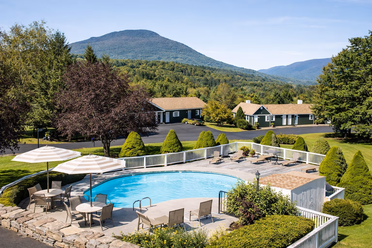 Outdoor pool with lounge chairs, umbrellas, and tables surrounded by greenery and cottages with mountains beyond.