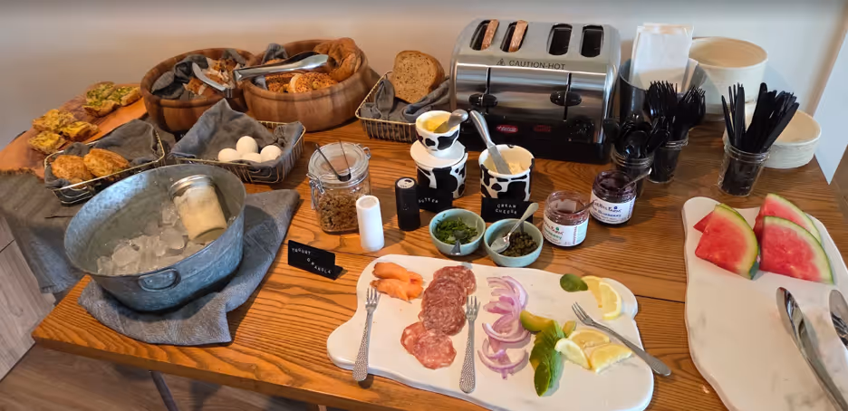 Breakfast spread with breads, sliced meats, onions, lemons, yogurt, jams, watermelon, and a toaster.