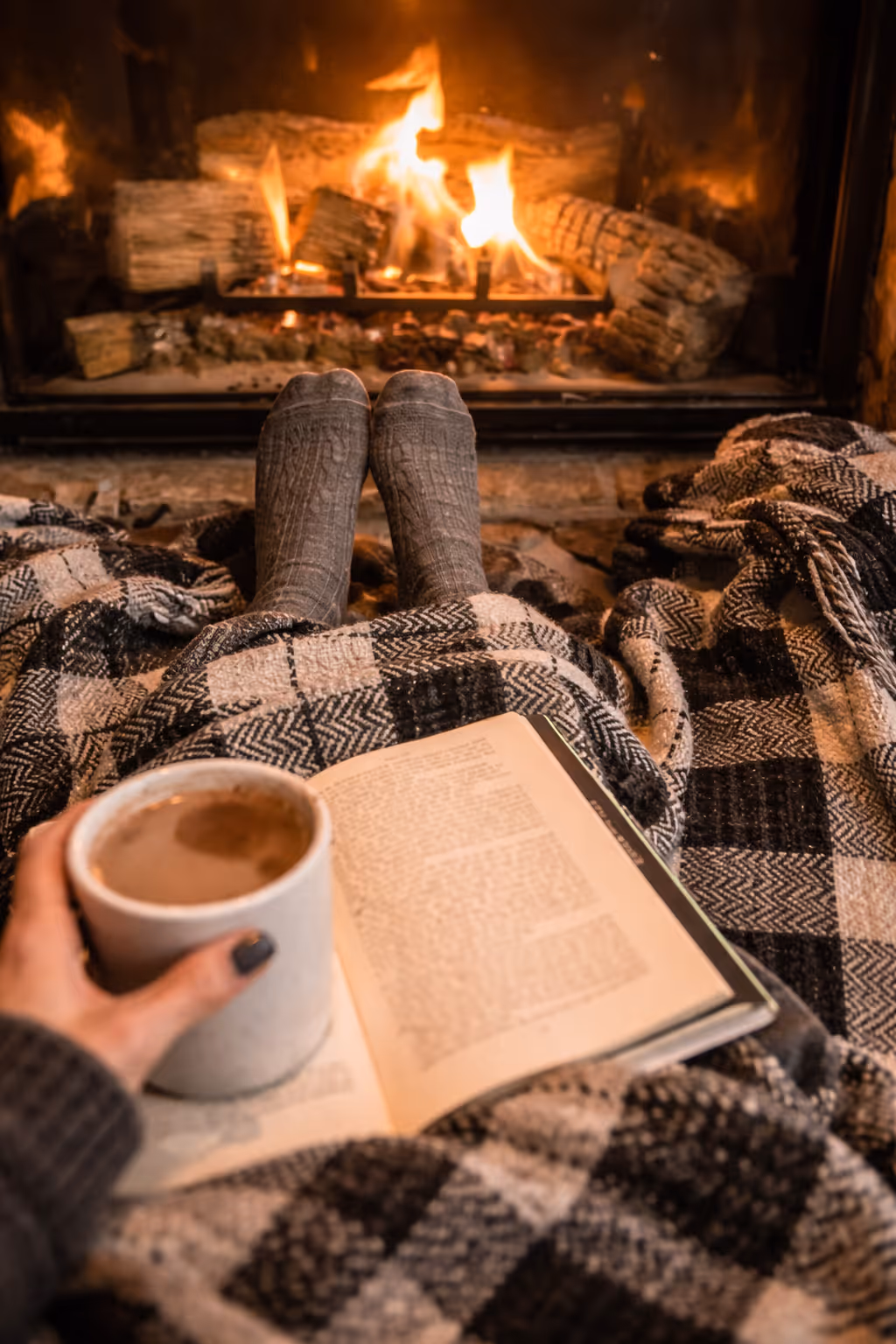 Person with gray socks under plaid blanket holding mug and book near a cozy fireplace.
