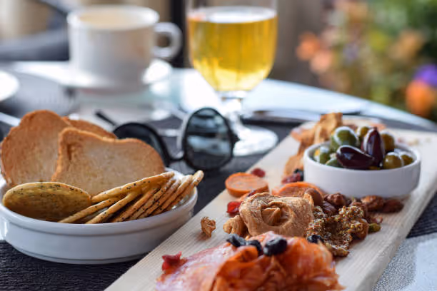 Charcuterie board with smoked salmon, dried fruit, nuts, olives, and crackers with toast and drinks in background.