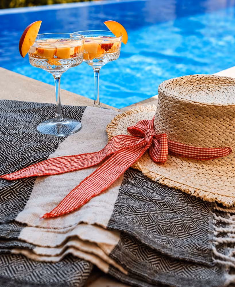 Two fruit cocktails garnished with peach slices next to a straw hat by the poolside.