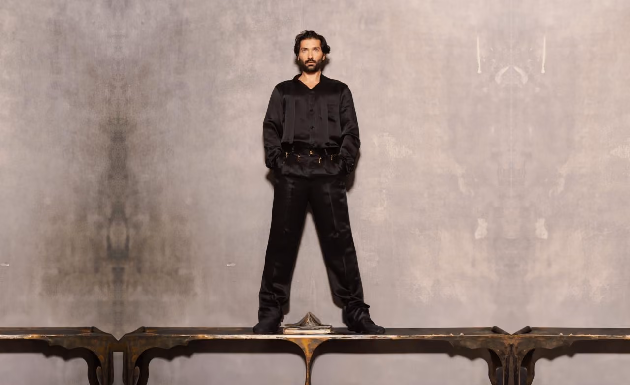 Man in elegant black suit standing on an antique wooden table against grey wall.