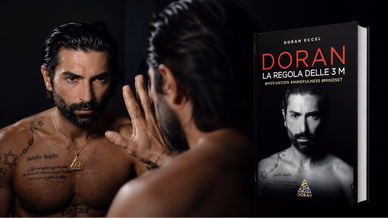Doran with tattoos and necklace looks at his reflection while next to him is a book by Doran about 3 m.