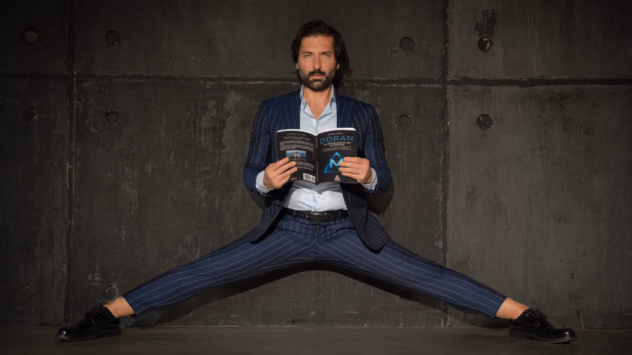 Man in elegant suit doing a split and reading a book on a dark wall background.