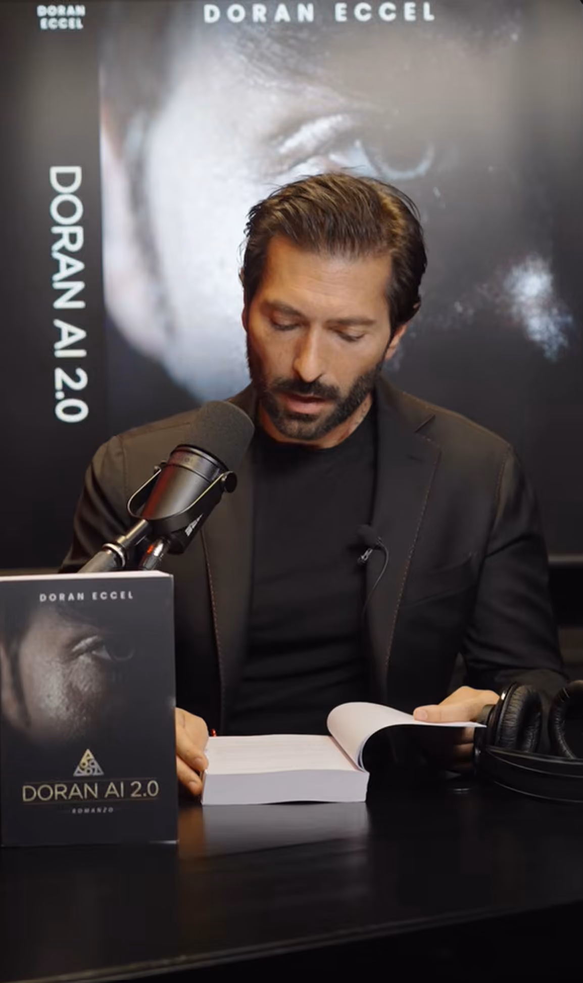 Man in a black jacket reads a book in front of a microphone, with a 'DORAN AI 2.0' book and headphones on his desk.