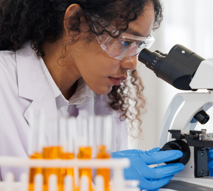 Scientist wearing protective goggles and blue gloves looking through a microscope with test tubes in the foreground.