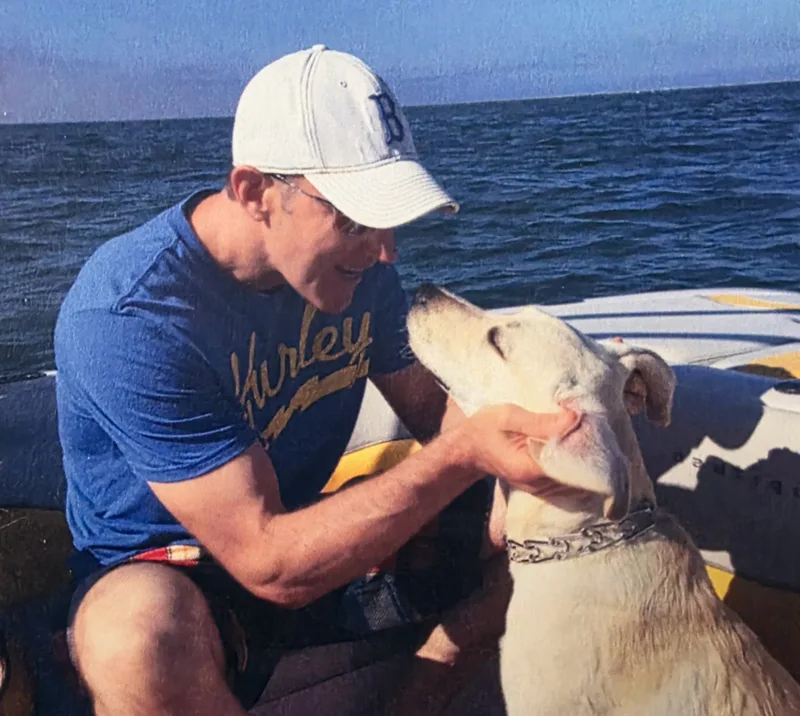 Vetabolics founder Rick Galyean wearing a white cap and blue shirt kneeling and gently holding his light-colored dog by the face near a body of water.