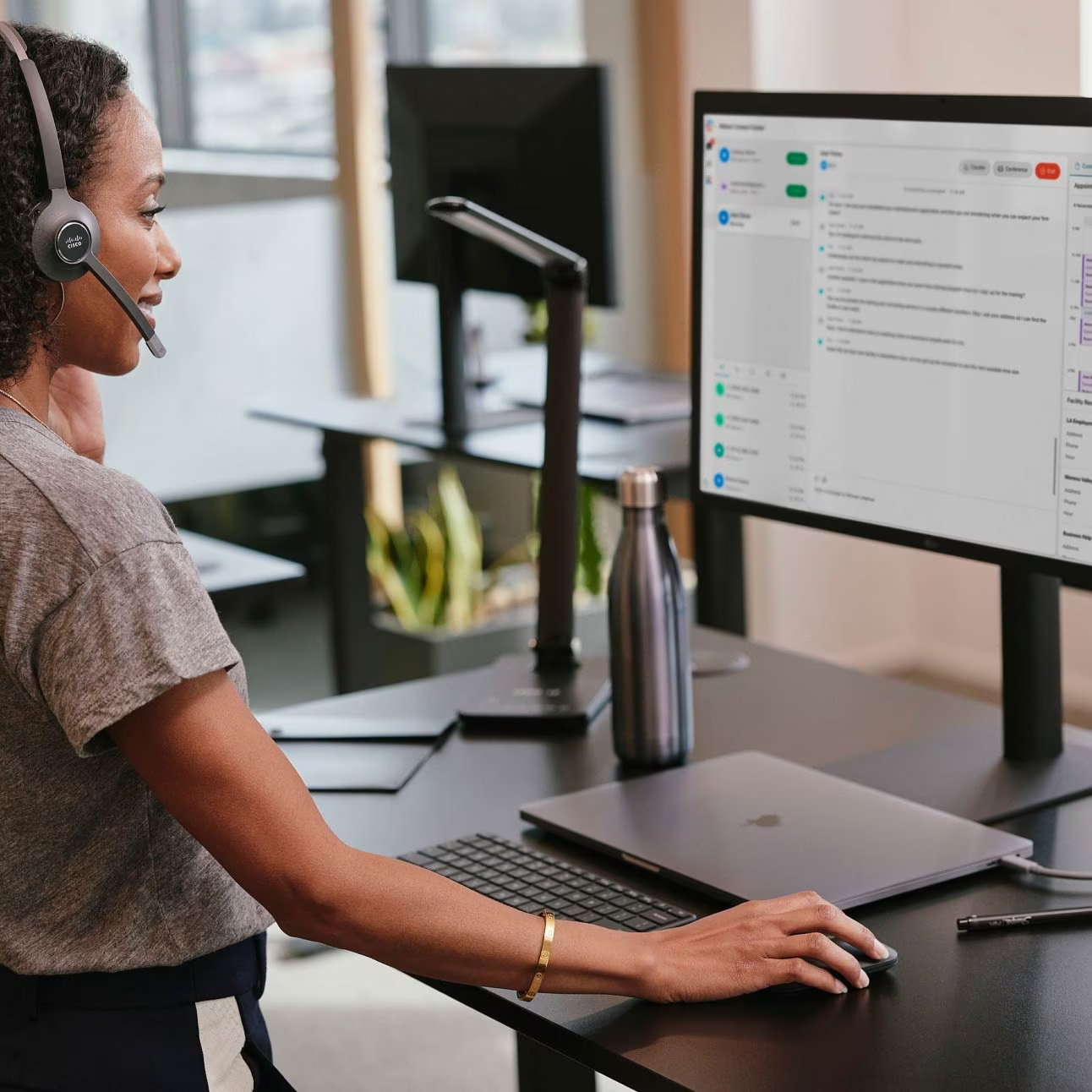 Woman wearing a headset using a computer mouse while looking at a monitor displaying a messaging application in a modern office.