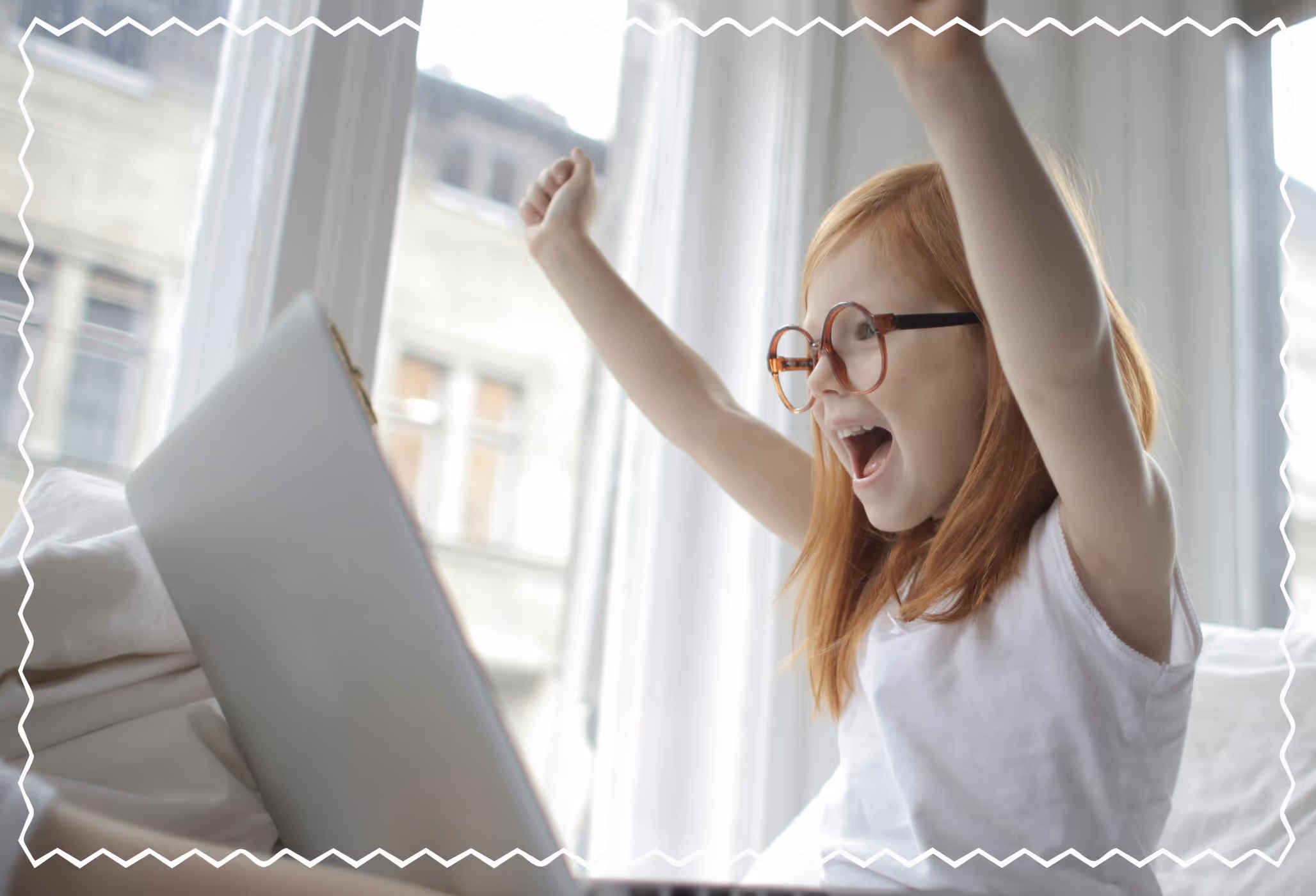 Excited young girl with red hair and glasses raising her arms while using a laptop at home.