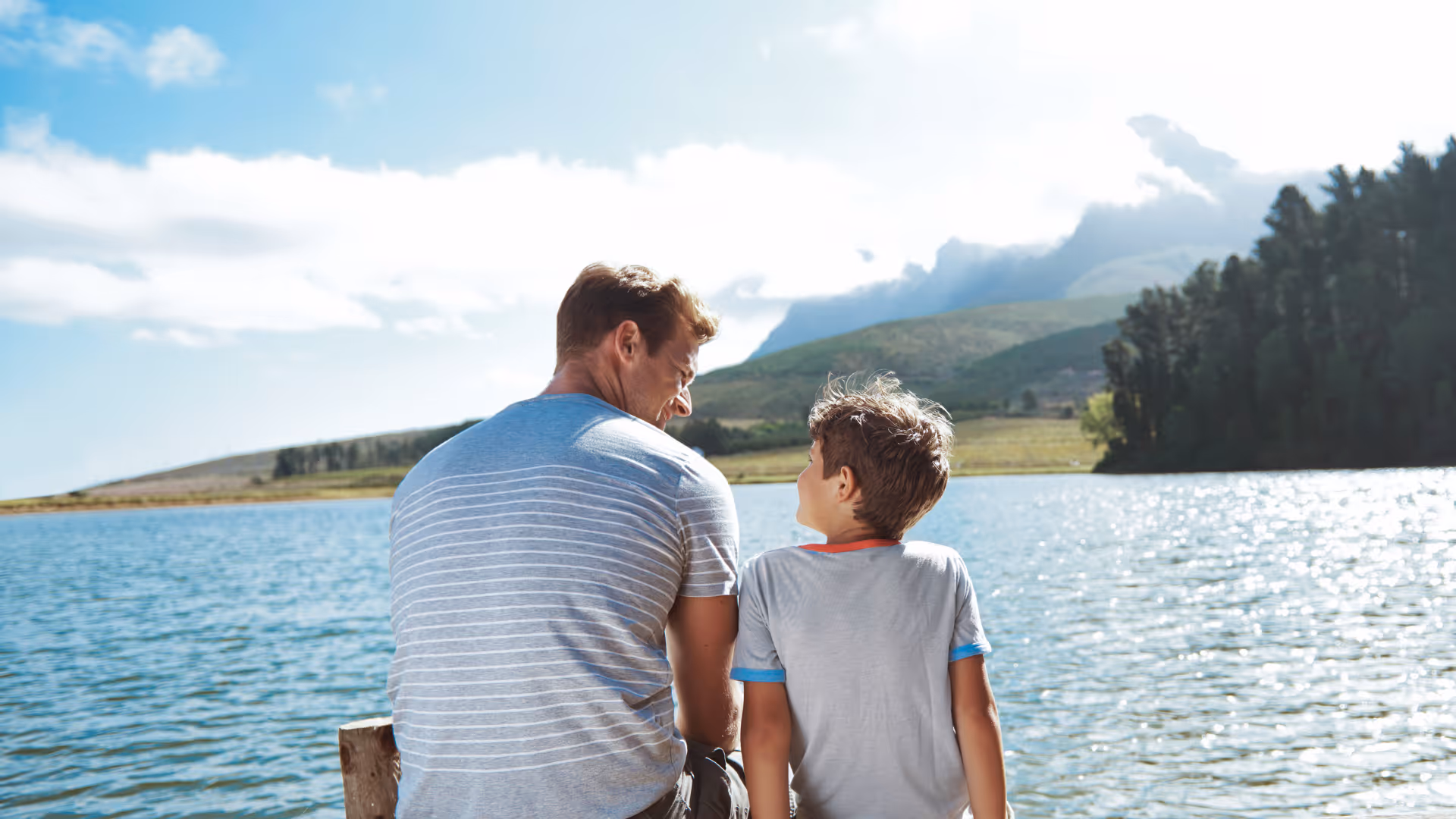 Man and boy sitting on a wooden dock beside a lake, looking at each other with mountains and trees in the background.