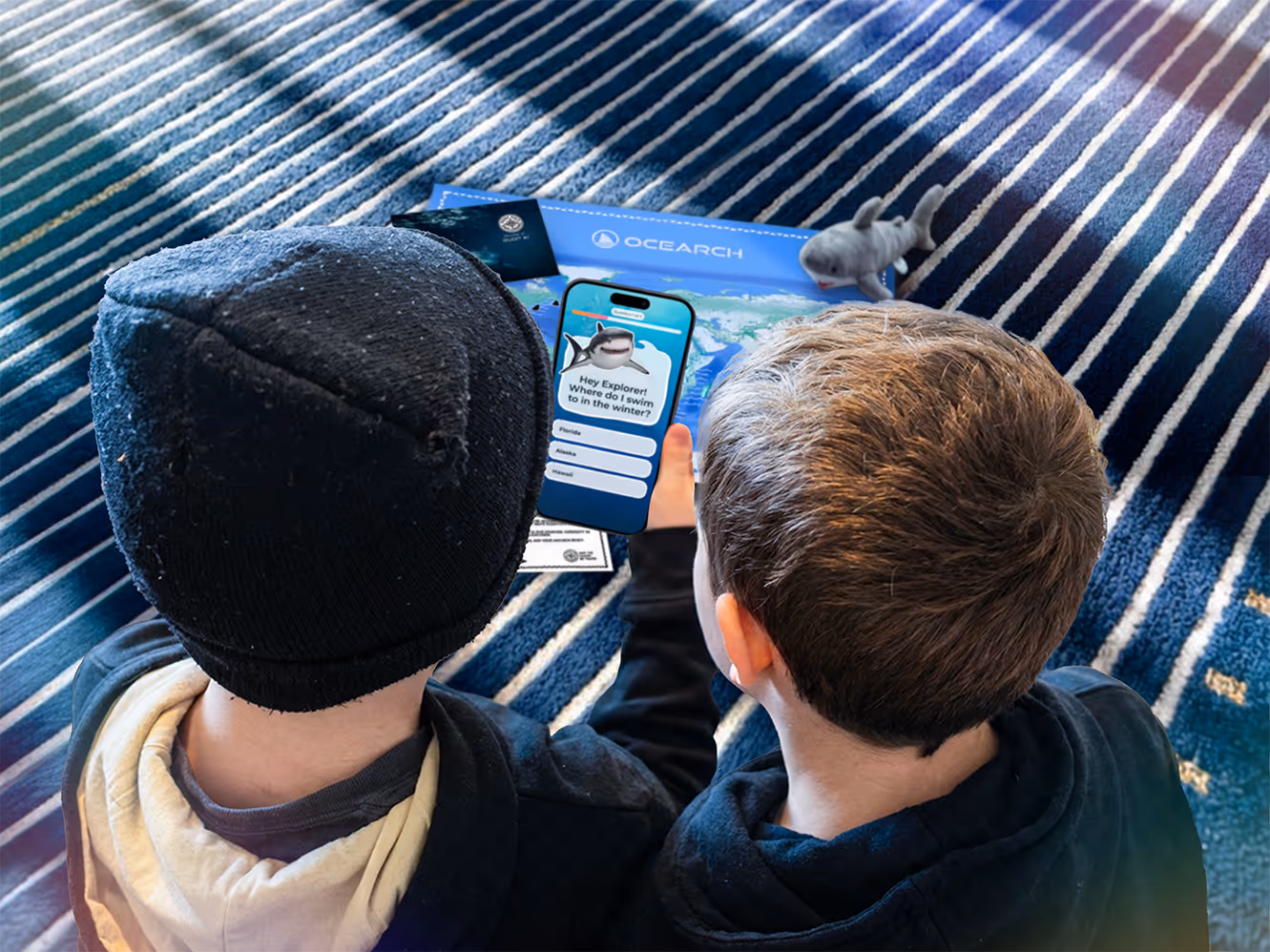 Two children sitting on a striped blue carpet looking at a phone showing a shark-themed quiz question from OCEARCH.