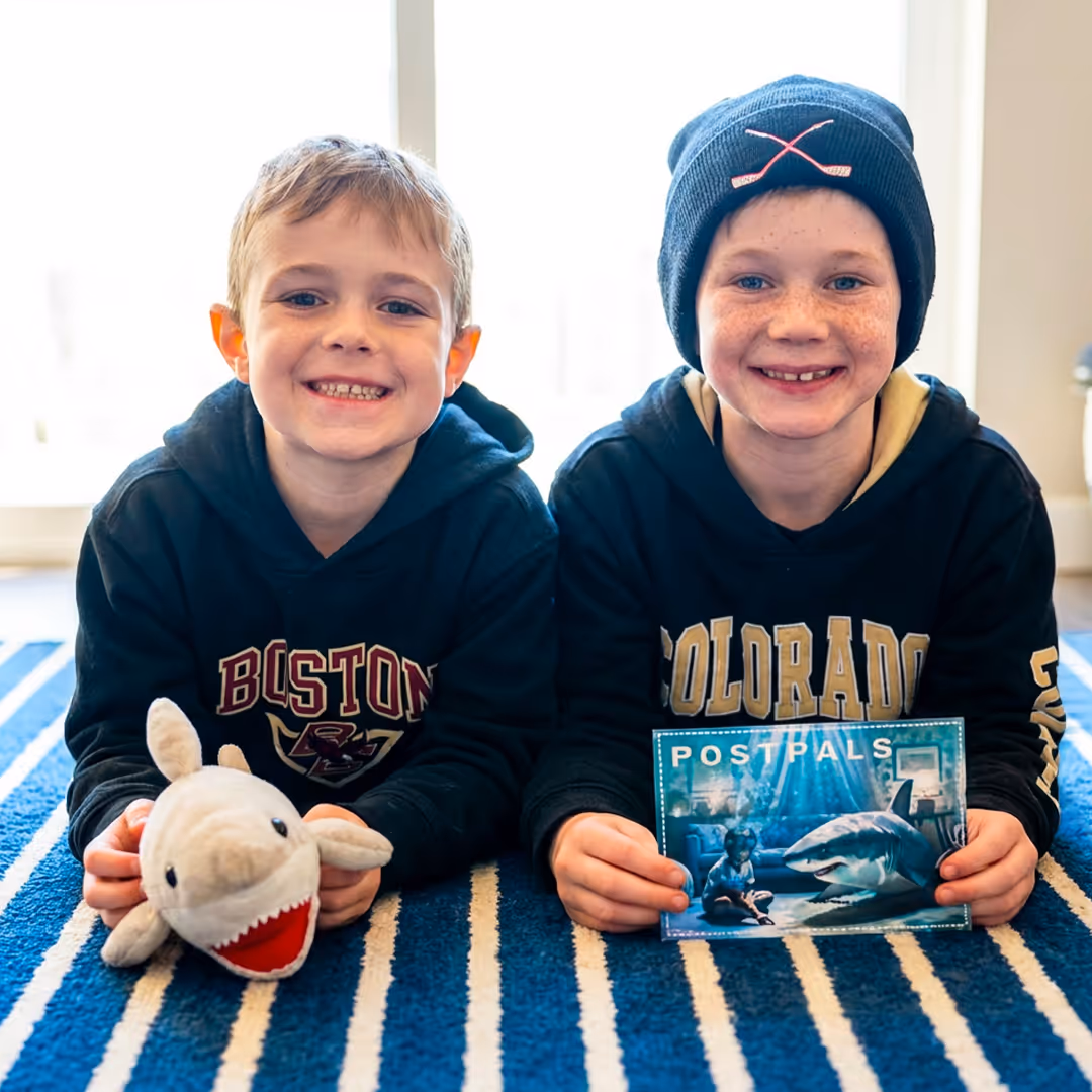 Two smiling boys lying on a blue and white striped carpet, one holding a plush shark and the other holding a postcard with a shark image.