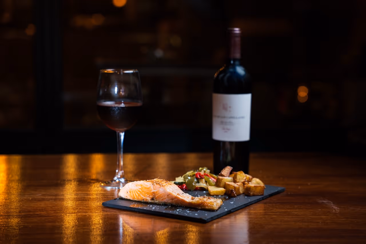 Filete de salmón asado con guarnición de papas y verduras acompañado de una copa y botella de vino tinto sobre mesa de madera.