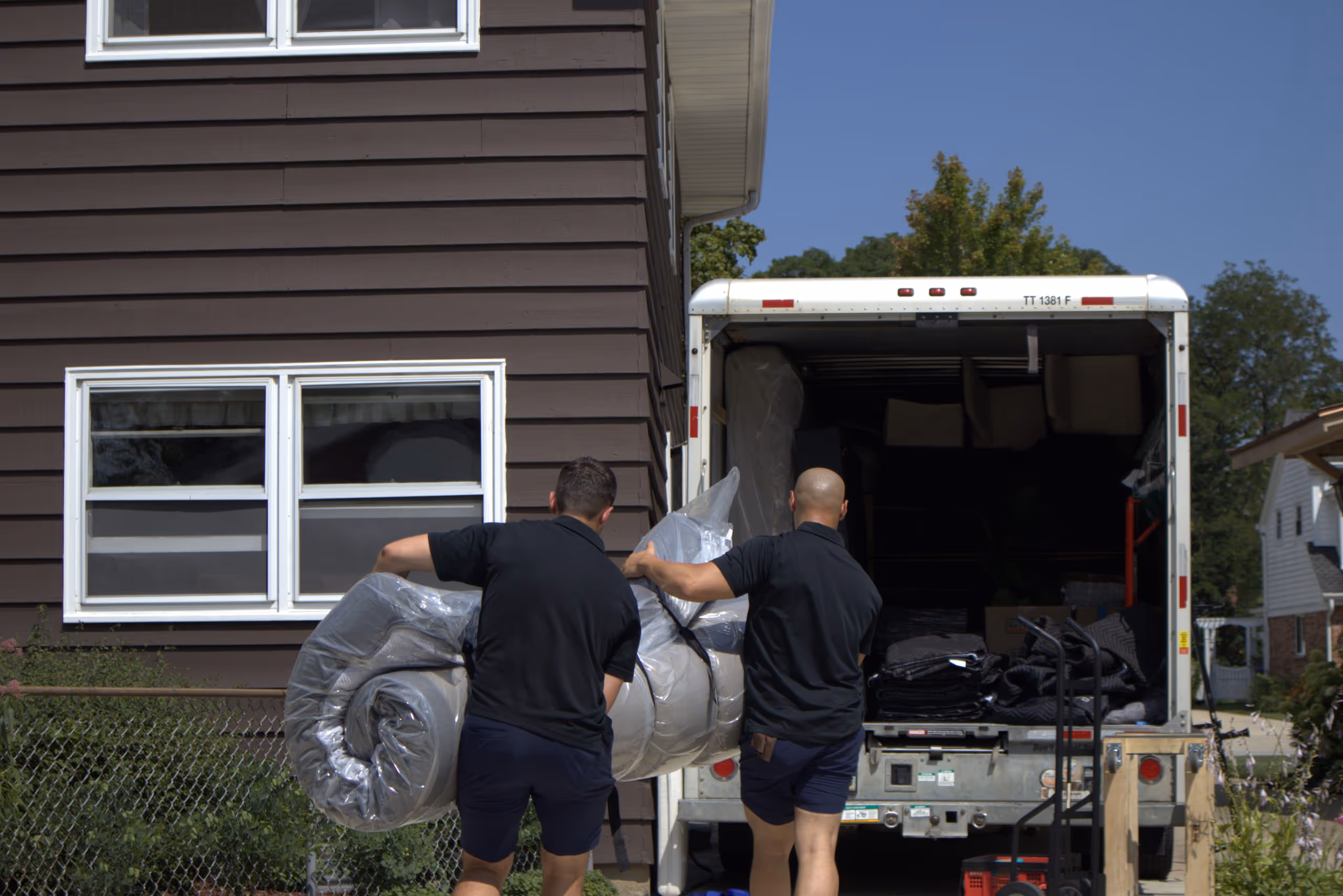 Two men in black shirts and shorts carrying a rolled mattress wrapped in plastic towards a moving truck in front of a brown house.