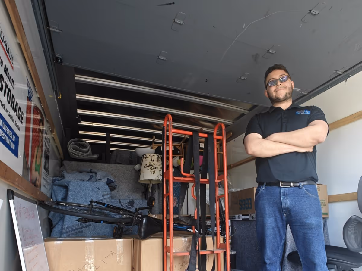 Man wearing glasses, black polo, and blue jeans standing inside a moving truck filled with boxes, a bicycle, and furniture.