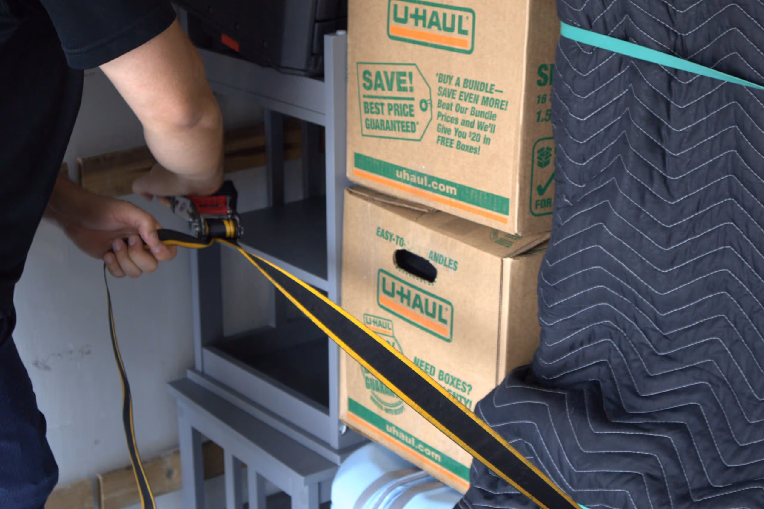 Person securing U-Haul moving boxes and furniture with a ratchet strap inside a moving truck.
