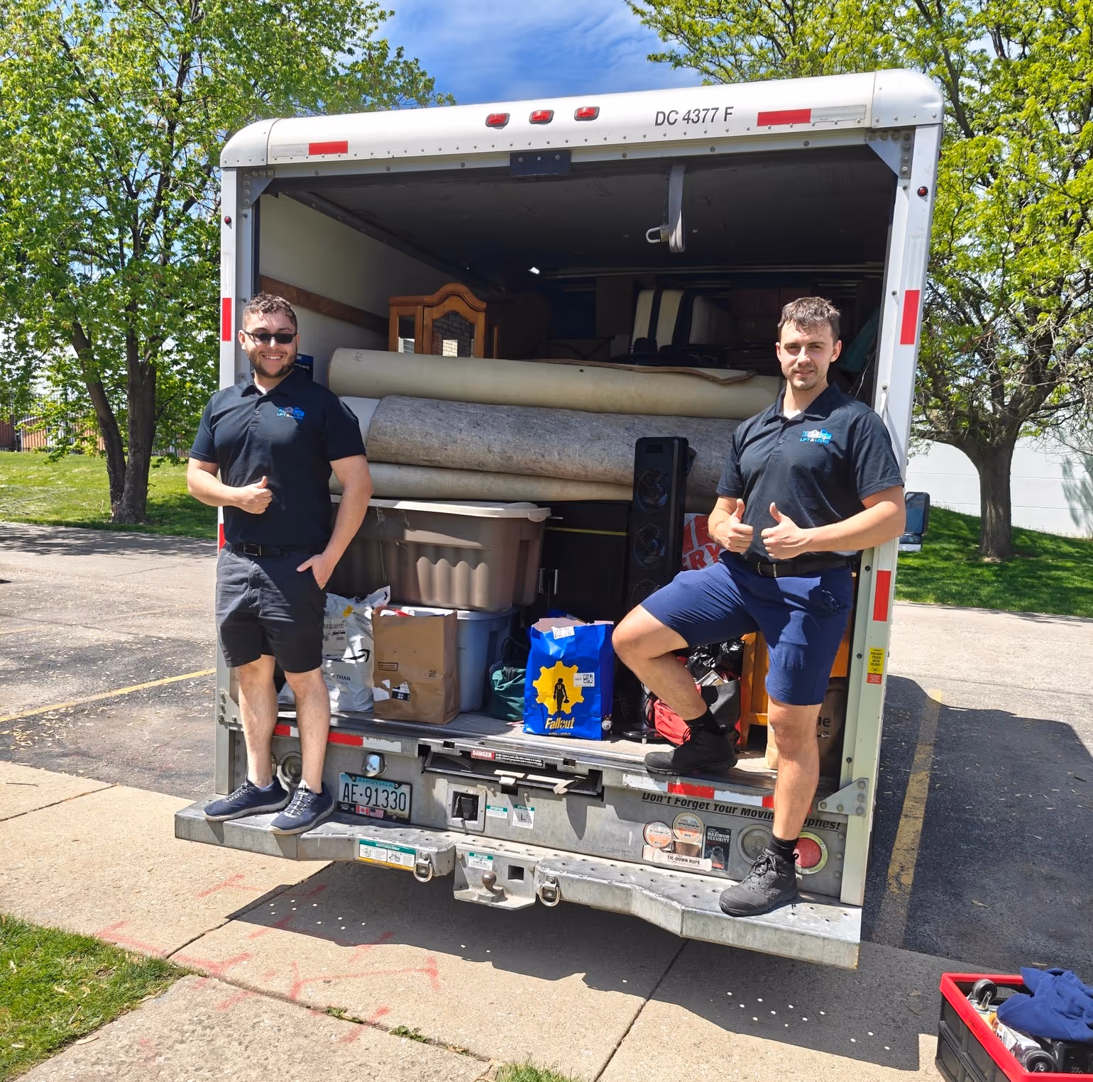 Two men wearing black polo shirts and shorts giving thumbs up while standing on the back bumper of a packed moving truck in an outdoor parking area.