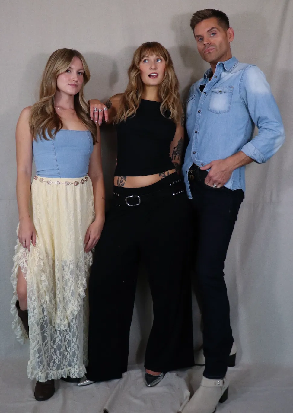 Three young adults posing against a plain backdrop: a woman in a blue top with a long lace skirt, a woman in a black sleeveless top and wide black pants, and a man in a light denim shirt with dark pants and white boots.