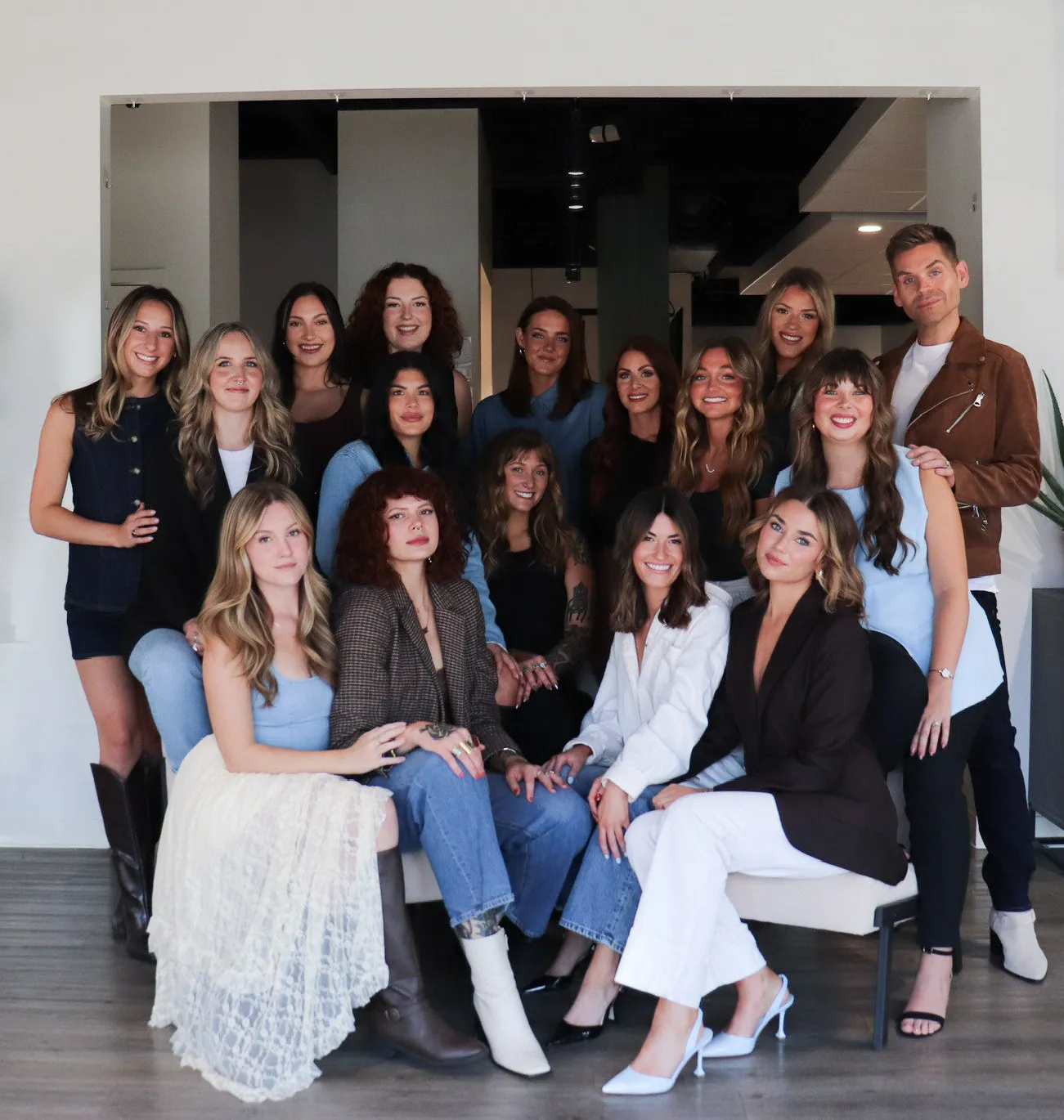 Group of fifteen smiling people posing indoors, with most seated and a few standing behind in casual and semi-formal attire.