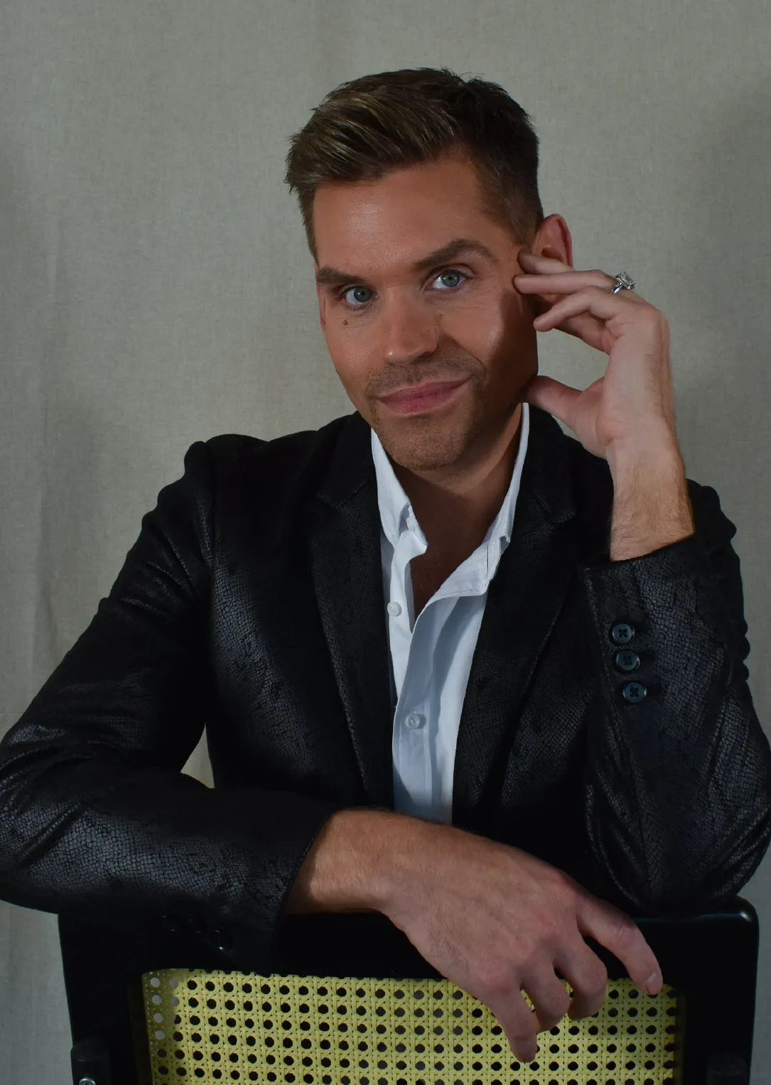 Man with short brown hair wearing a black blazer and white shirt, sitting on a chair and resting his hand on his face while smiling slightly.