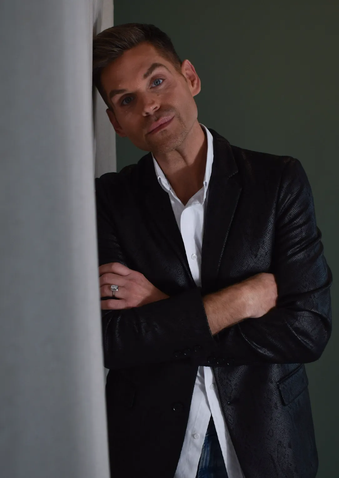 Chris, Stylist at Sul The Studio, with short brown hair leaning against a wall, wearing a black textured blazer over a white shirt with arms crossed.
