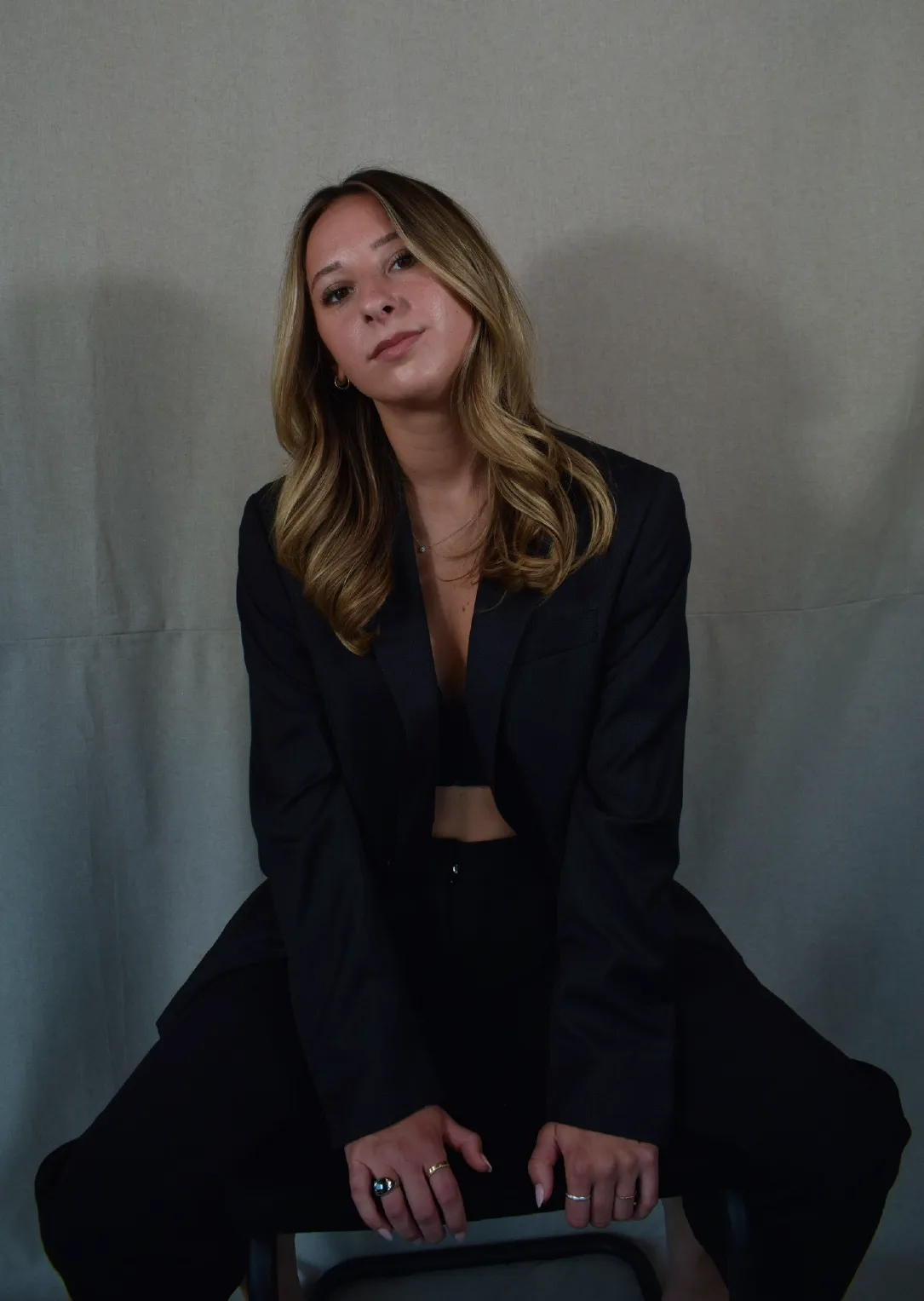 Young woman with long blonde hair wearing a black blazer and pants, sitting on a chair against a plain gray backdrop.