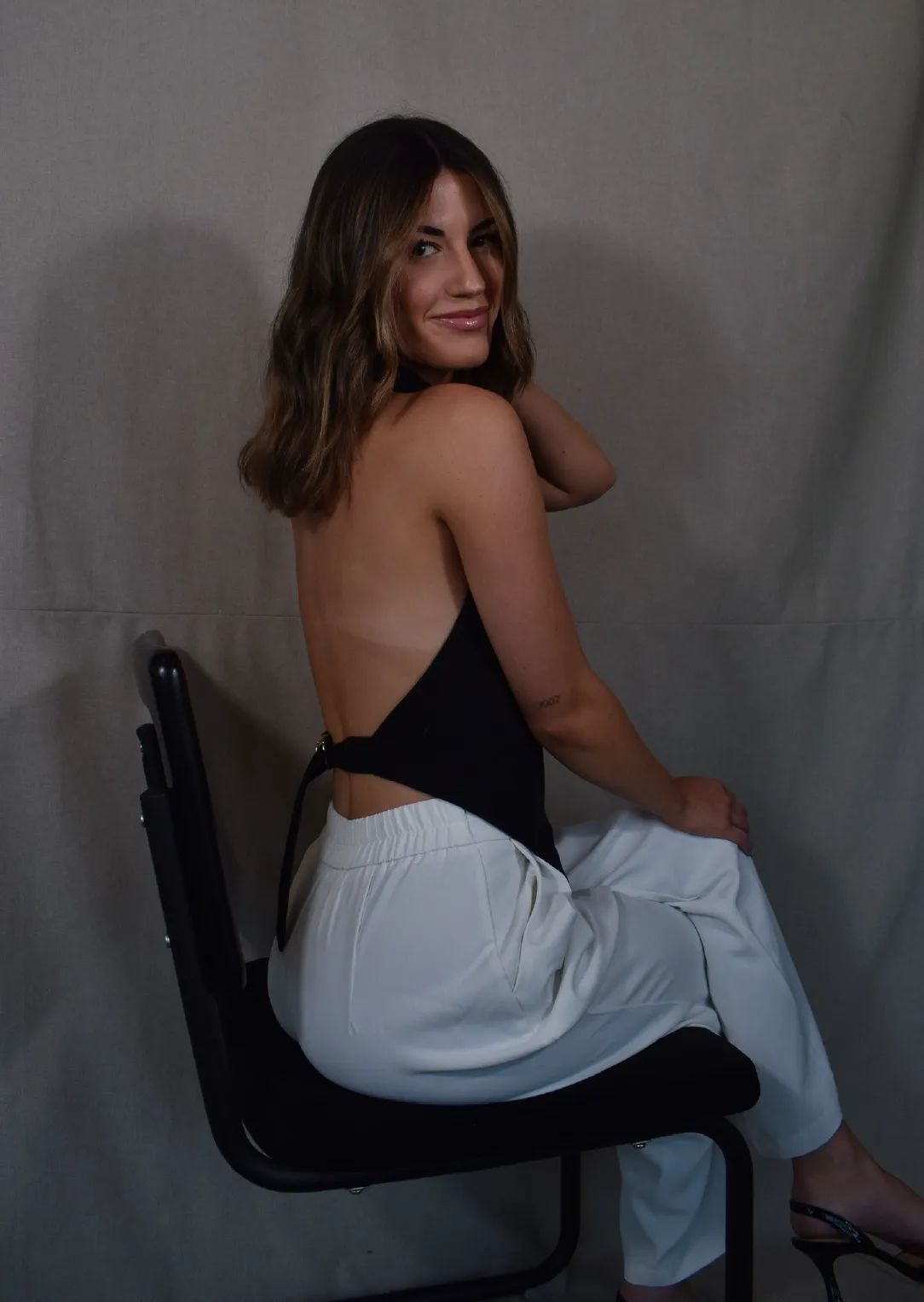 Woman with shoulder-length wavy hair wearing a backless black top and white pants sitting sideways on a black chair, smiling at the camera against a gray backdrop.