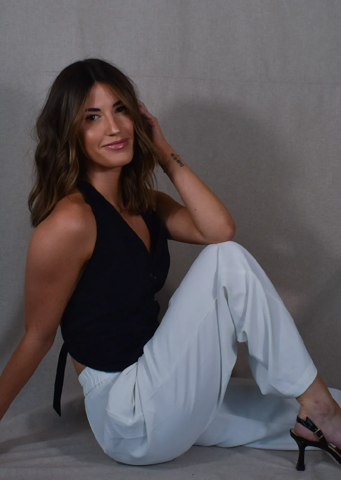 Woman with shoulder-length wavy brown hair sitting on the floor, wearing a black sleeveless top, white pants, and black heeled sandals.