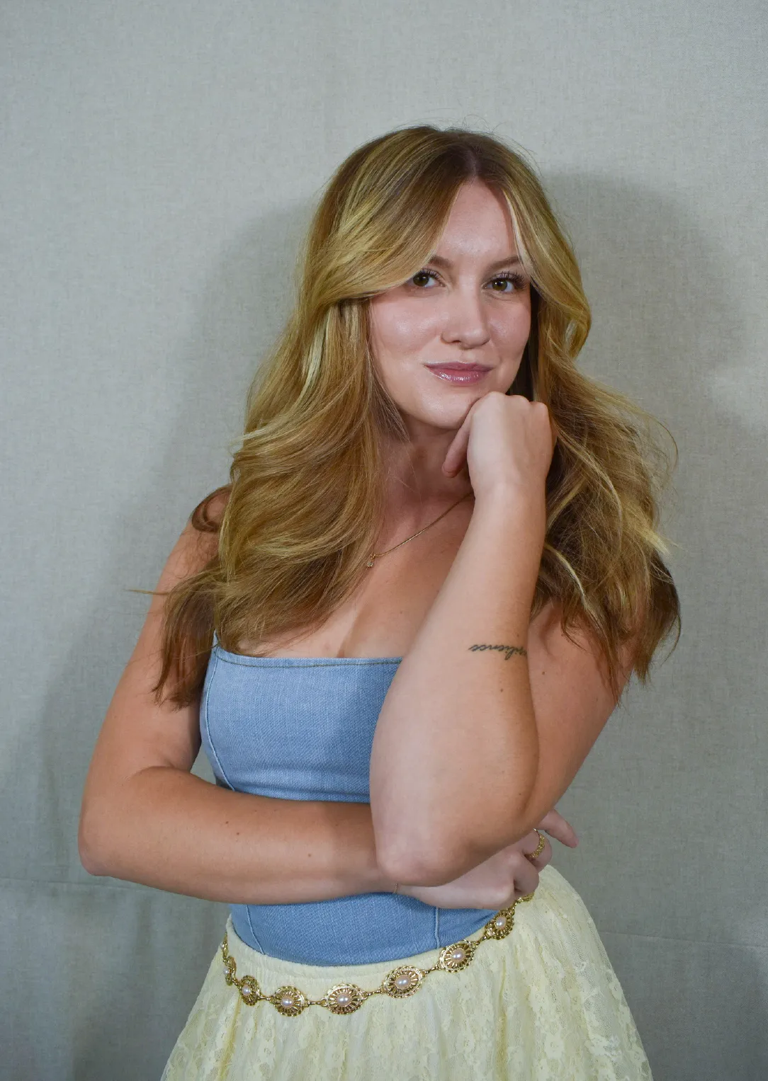 Nayla, Stylist at Sul The Studio, with long blonde hair wearing a blue strapless top and yellow lace skirt with a decorative belt, posing with hand on chin.