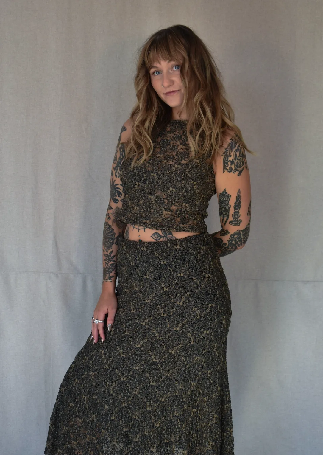 Sam, Stylist at Sul The Studio, with wavy light brown hair and multiple tattoos on her arms and torso wearing a dark patterned sleeveless top and matching long skirt, posing in front of a gray backdrop.
