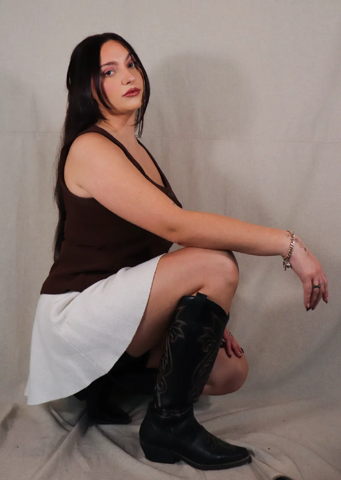 Alyssa, Stylist at Sul The Studio, with long dark hair wearing a brown sleeveless top, white skirt, and black cowboy boots, squatting against a plain beige background.