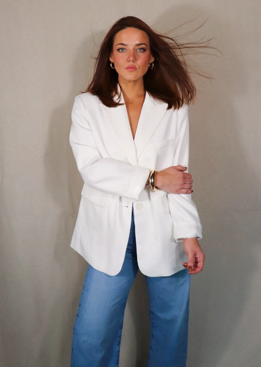Claire, Stylist at Sul The Studio, with shoulder-length brown hair wearing a white blazer and blue jeans, standing against a neutral background.