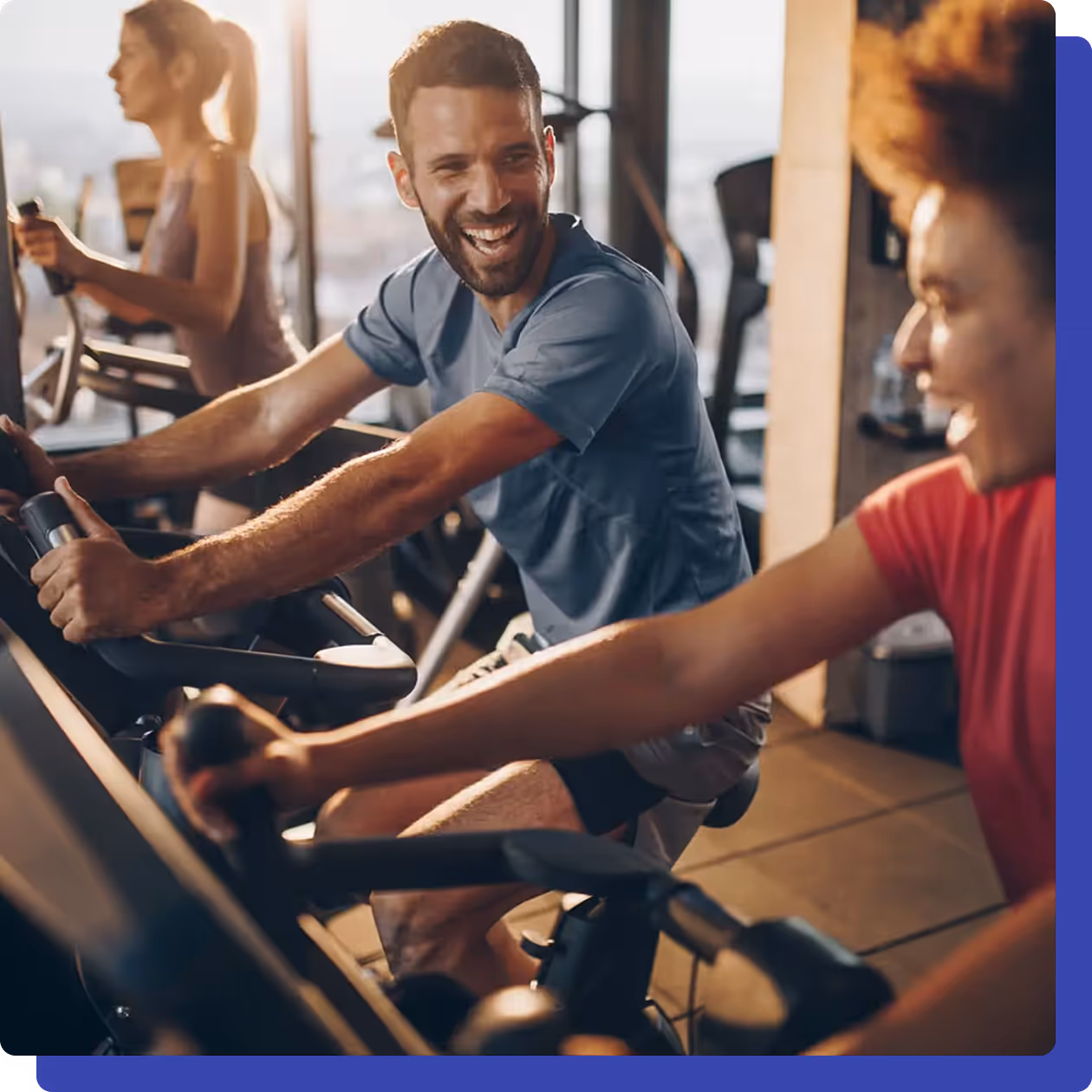 Smiling man and woman exercising on stationary bikes at the gym, enjoying a workout together with others in the background.