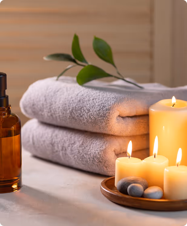 Spa setting with folded towels, burning candles, massage stones, and an amber glass bottle on a relaxing surface.