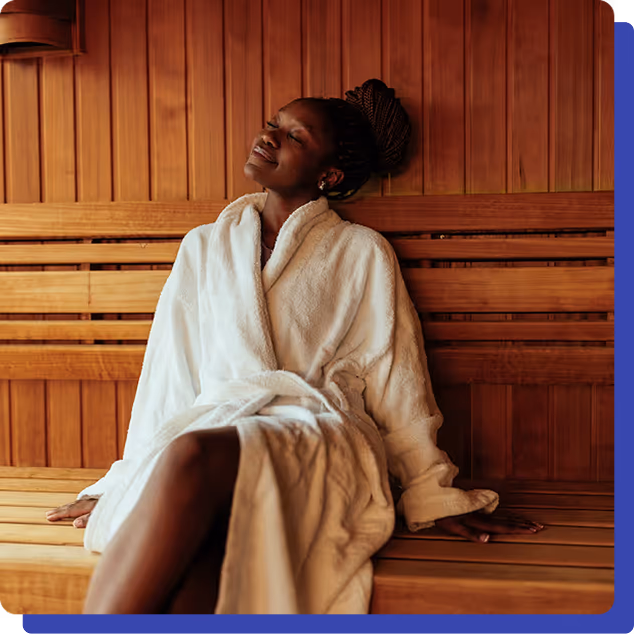 Relaxed woman in a white robe sitting in a wooden sauna, leaning back with eyes closed and enjoying the warmth.