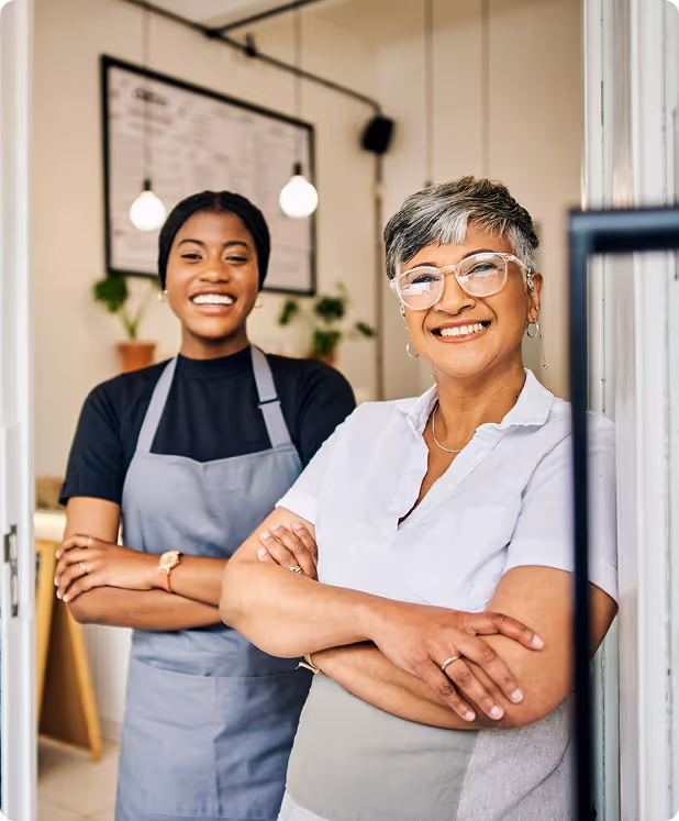 Two smiling women standing with arms crossed in a café entrance, wearing aprons and looking confident.