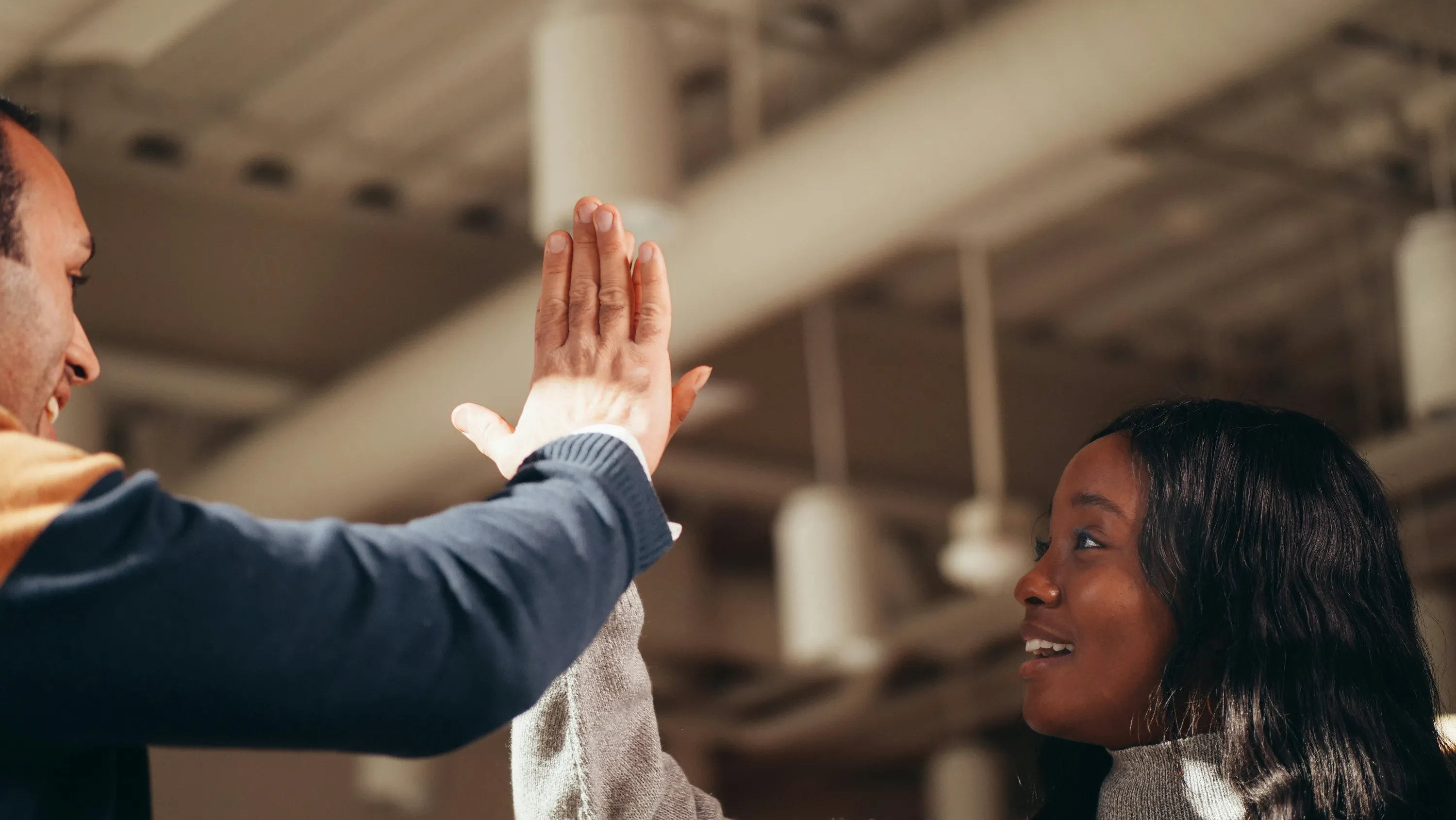 Two people smiling and giving a high five indoors with blurred industrial ceiling in background.