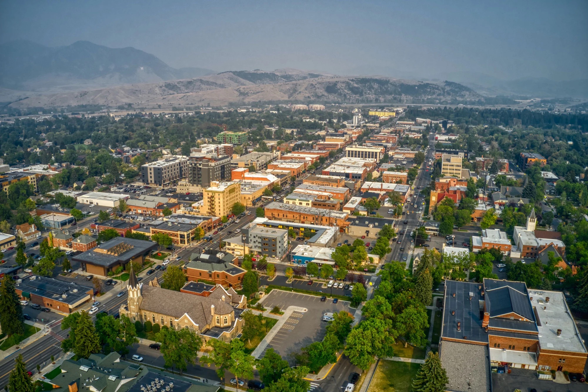 Aerial view of a city with tree-lined streets, low-rise buildings, and distant mountains under a clear sky.
