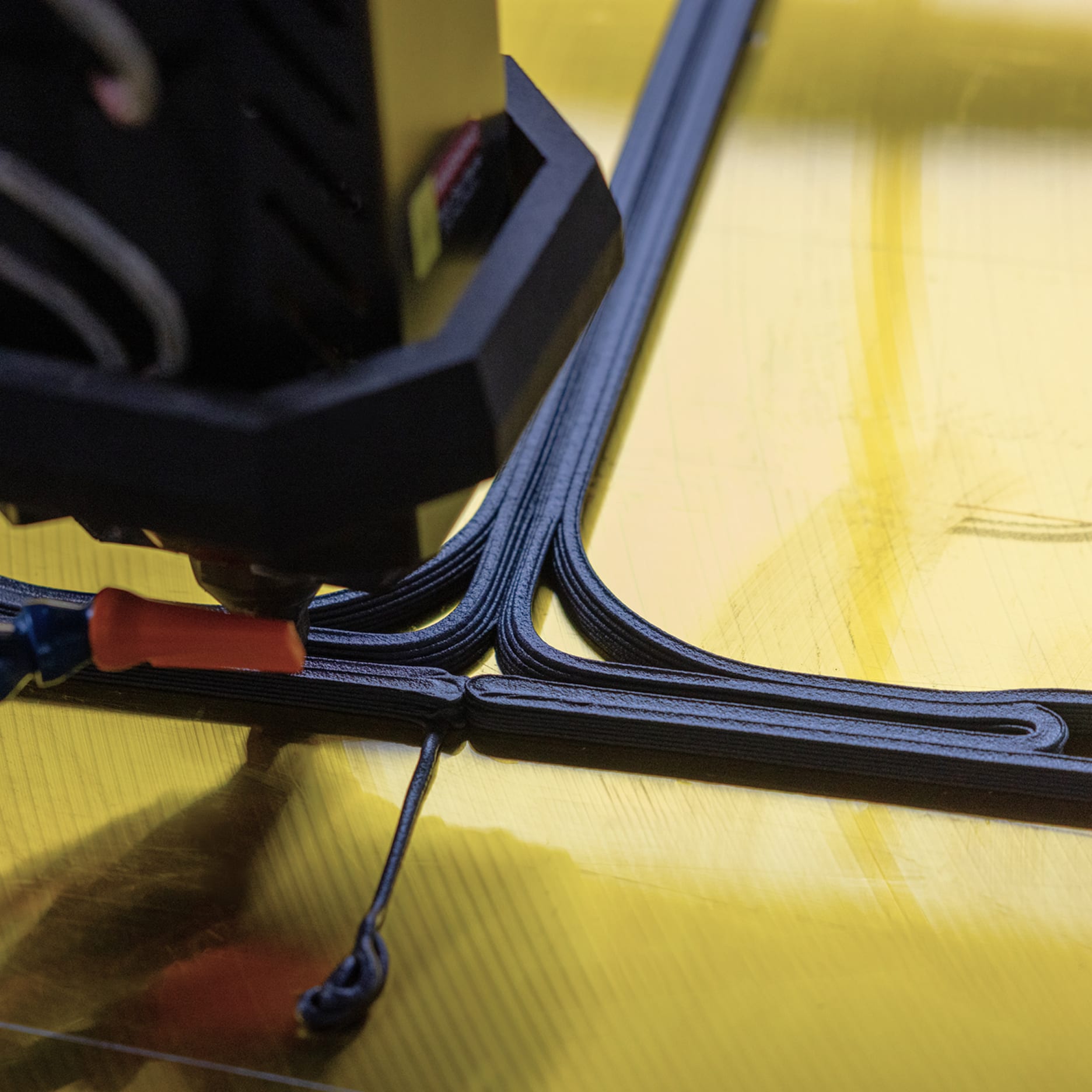 Close-up of a 3D printer extruding black filament in precise intersecting lines on a yellow surface.