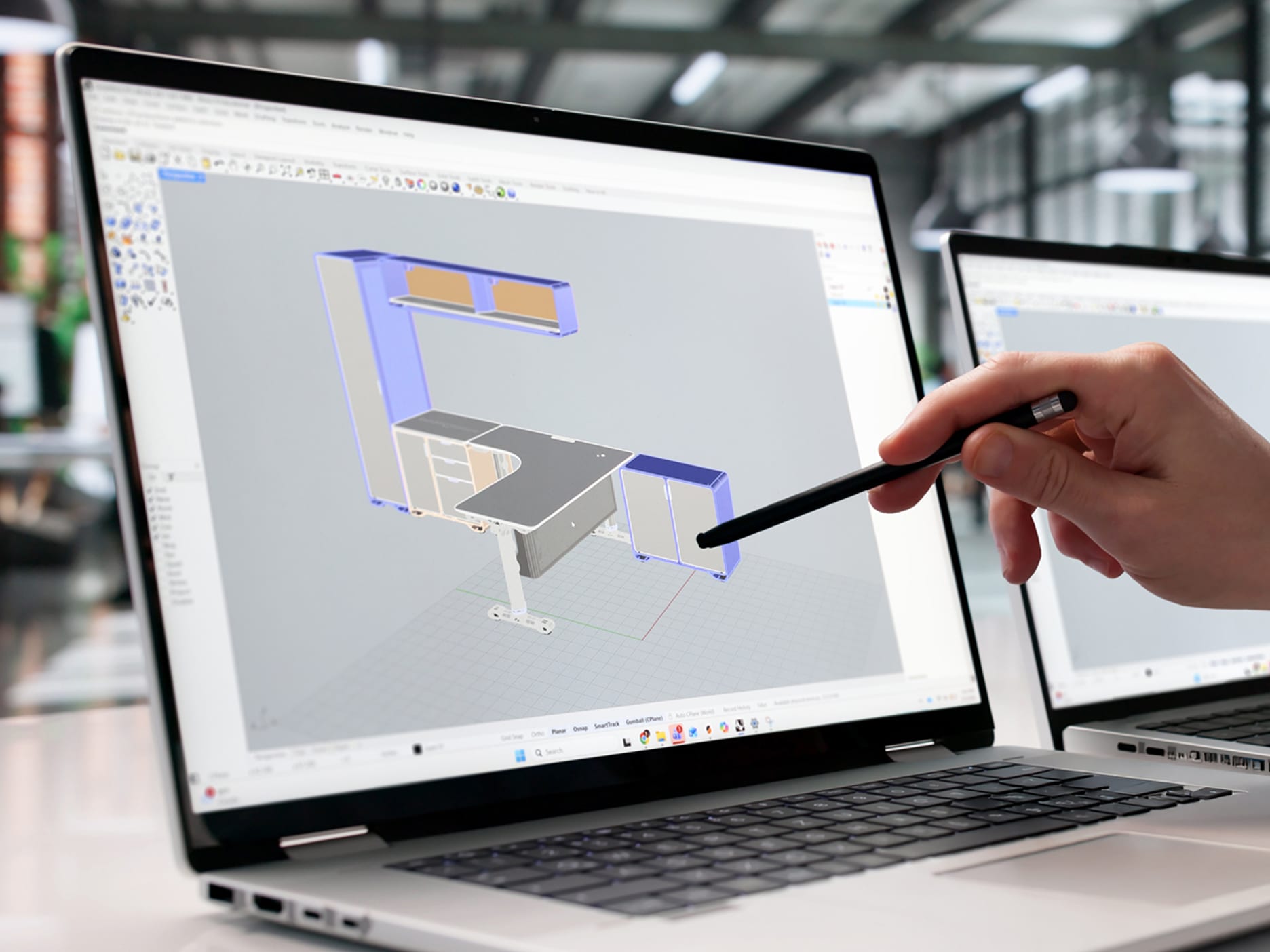 Laptop screen displaying a 3D design of a modular office desk setup with cabinets, while a hand points at the screen using a stylus pen.