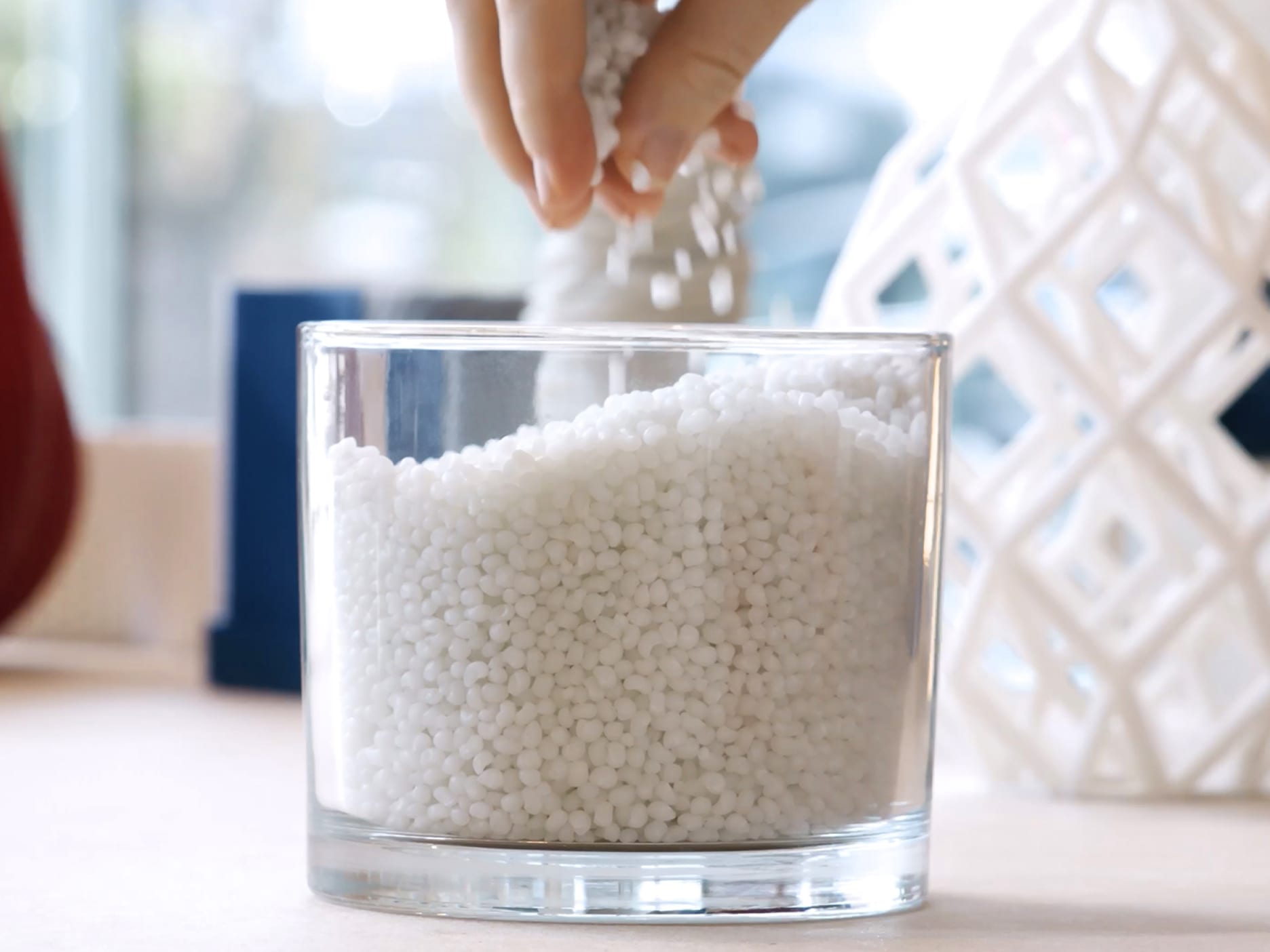 Hand pouring small white plastic pellets into a clear glass container on a table.