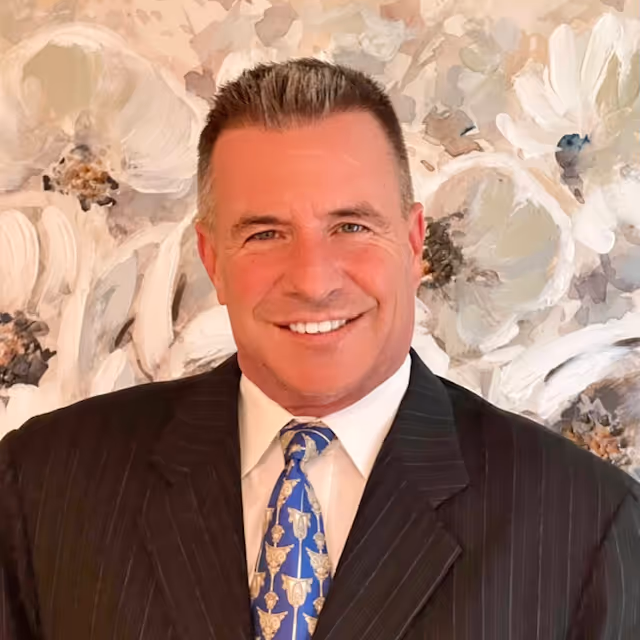 Smiling man in a dark pinstripe suit and blue patterned tie against a floral background.